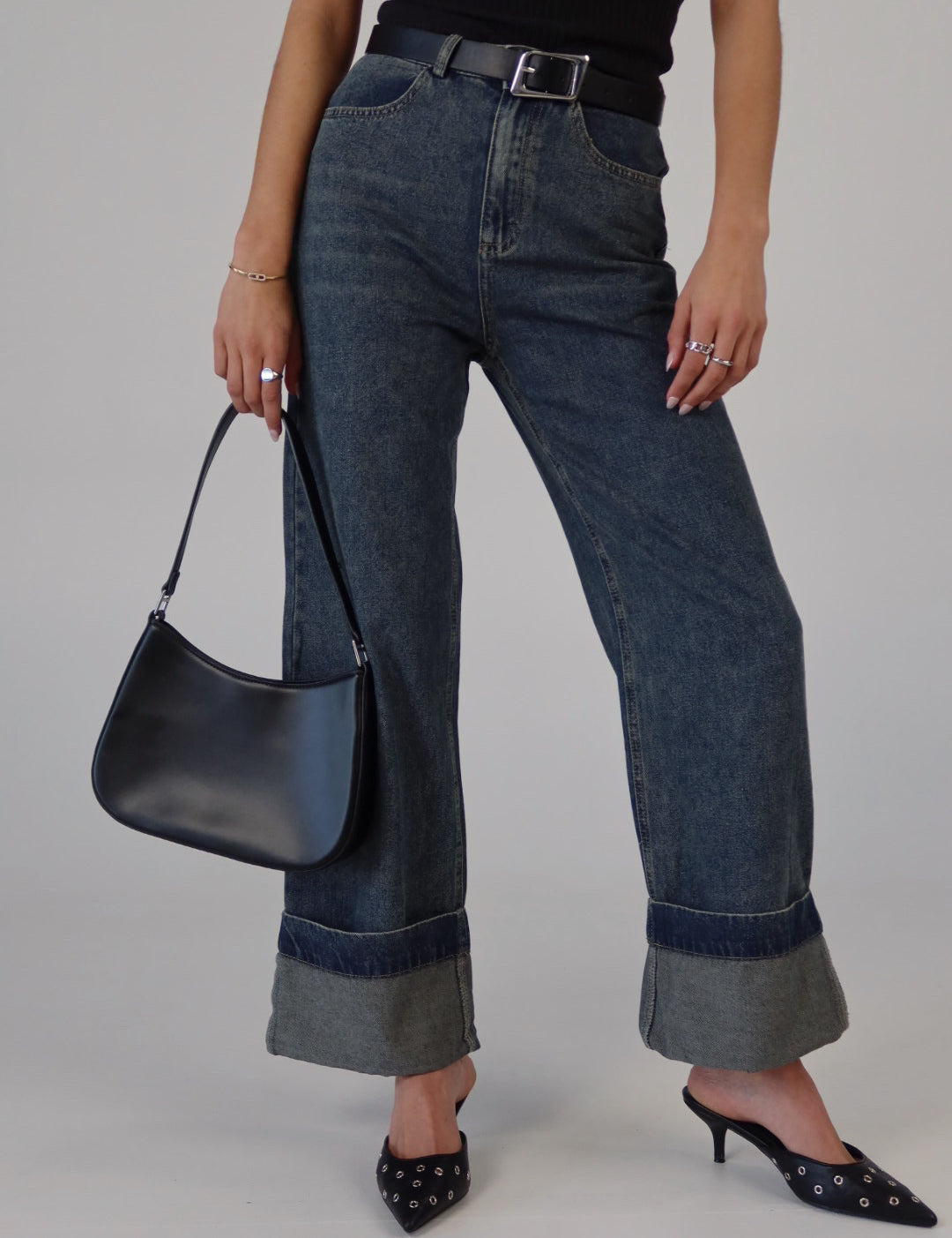 A person standing wearing fold-over washed denim pants with a black belt, black bag, and black shoes.