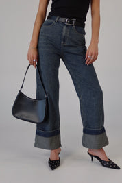A person standing wearing fold-over washed denim pants with a black belt, black bag, and black shoes.